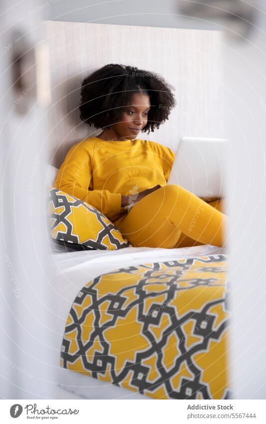 Black woman in digital moment at home yellow laptop bed cushion room light interior attire technology relax study browse female online comfort bedroom casual