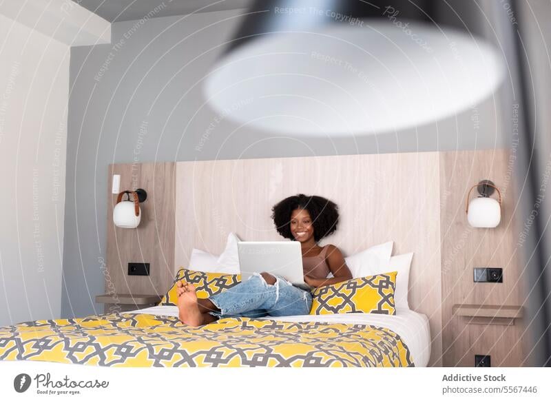 Black woman in modern relaxation curly laptop bed decor cheerful work ethnic technology indoor panel wall lamp female young pillow pattern comfort