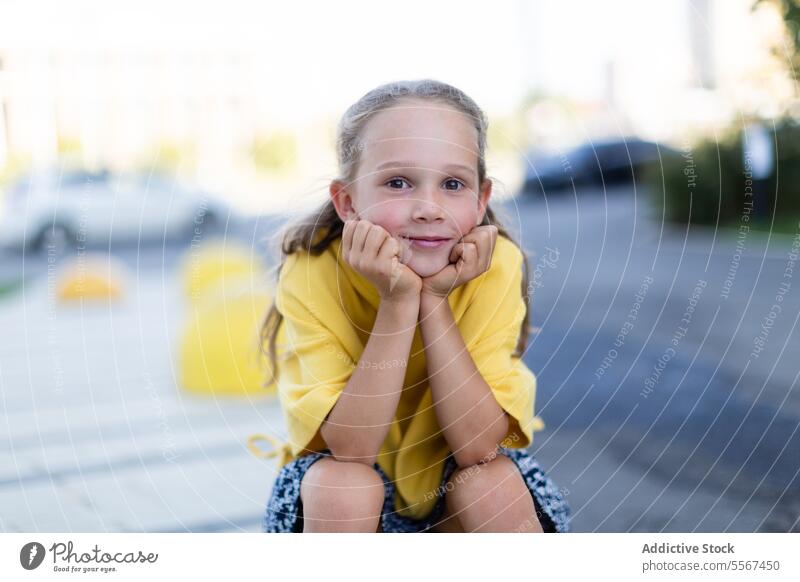 Radiant girl in yellow top blonde smile urban joy curb seat hand chin bright street child youthful radiant kid sit happy outdoor city casual expression genuine