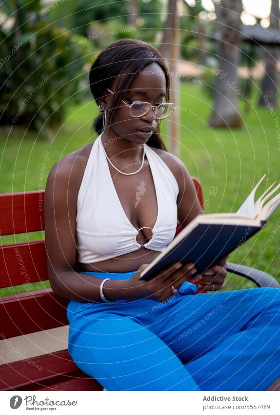 African American woman reading in garden book focus eyeglasses bench outdoors green relaxed halter white blue pants trees contemplative studious nature serenity