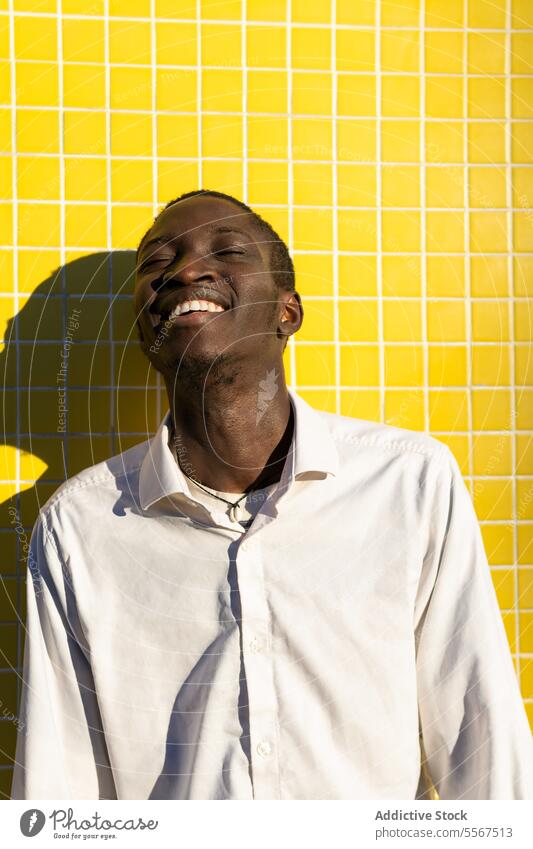 Young man with in Bright laughter young joyful yellow tile wall sunlight shadow white shirt bright vibrant happy cheerful positive emotion face teeth smile mood