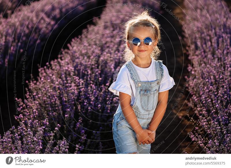 adorable child girl in lavender field on sunset. smiling kid in sunglasses, jeans jumpsuit is having fun on nature on summer day. Family day, vacation, holiday.International Children's Day,