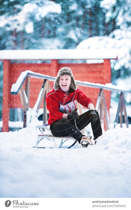 Smiling young cheerful caucasian man is wearing worm red sweatshirt and fun winter ear hat is having fun with sled in a snowy park or forest on winter day. Happy Christmas winter holiday