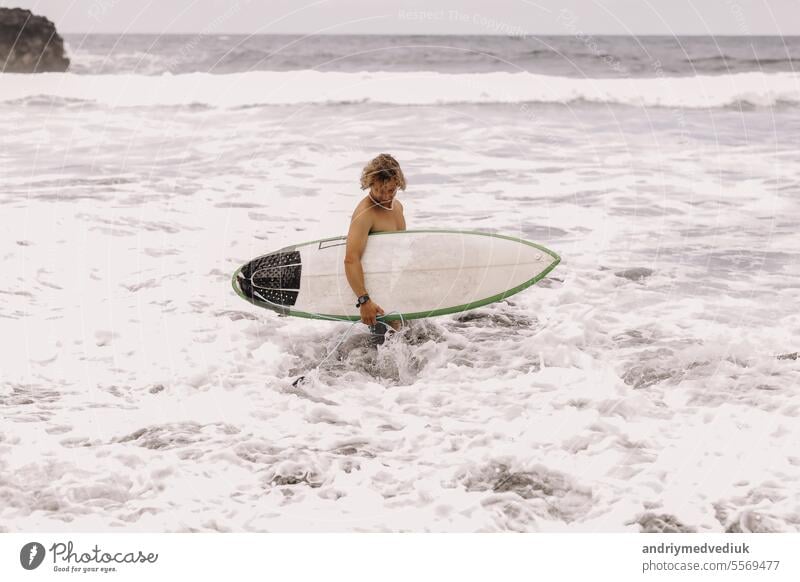 young fit surfer man with surfboard runs into ocean or sea with big waves for surfing. Concept of extreme sport, fitness, freedom, happiness, new modern life. Sports travel destination.