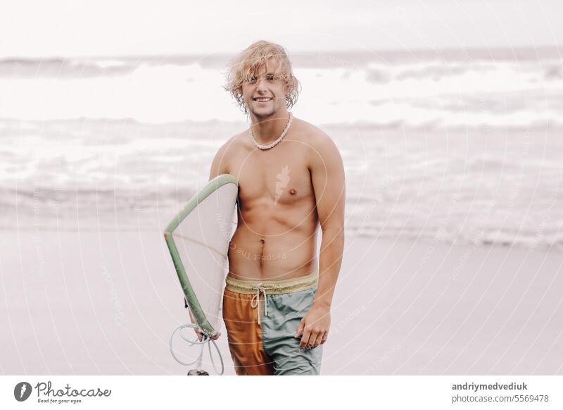 Handsome fit young blond man with mock up surfboard waits for wave to surf spot at sea ocean beach with black sand and looks at camera. Concept of sport, fitness, freedom, happiness, new modern life