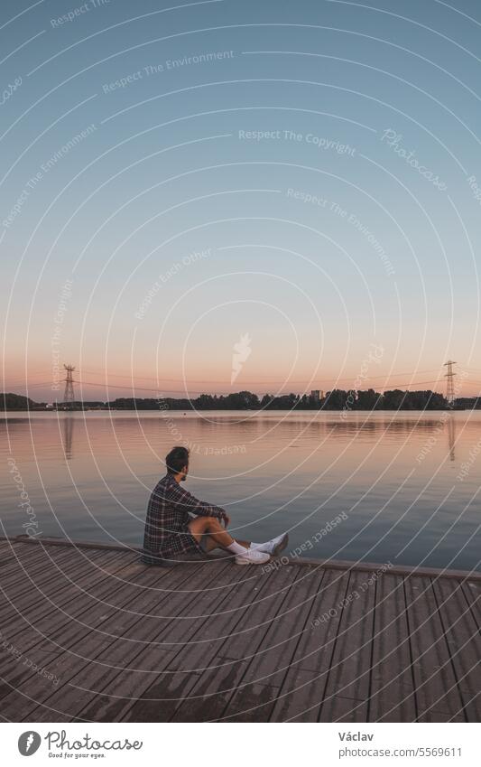 Young 25 year old brown man with plaid shirt sitting on the end of a wooden pier during sunset, watching the calm water surface in Almere Netherlands one