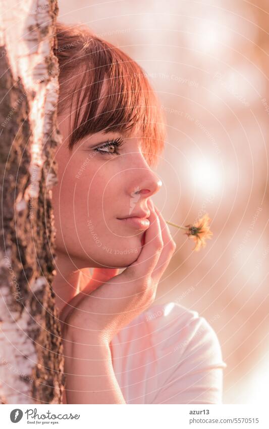Beautiful girl with flower dreaming in nature at tree trunk. Young carefree woman with best health at sunny summer day in youth time. Perfect skin with authentic natural beauty cosmetic makeup