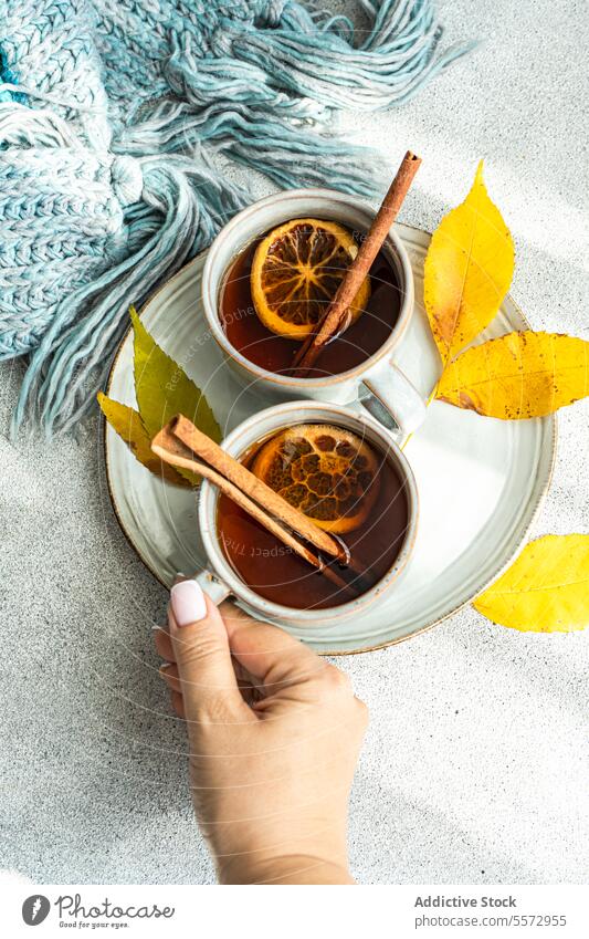 Unrecognizable hand reaching for autumn spiced tea cup cinnamon anise dried orange slice leaf yellow beverage blue scarf gray surface ceramic drink aromatic