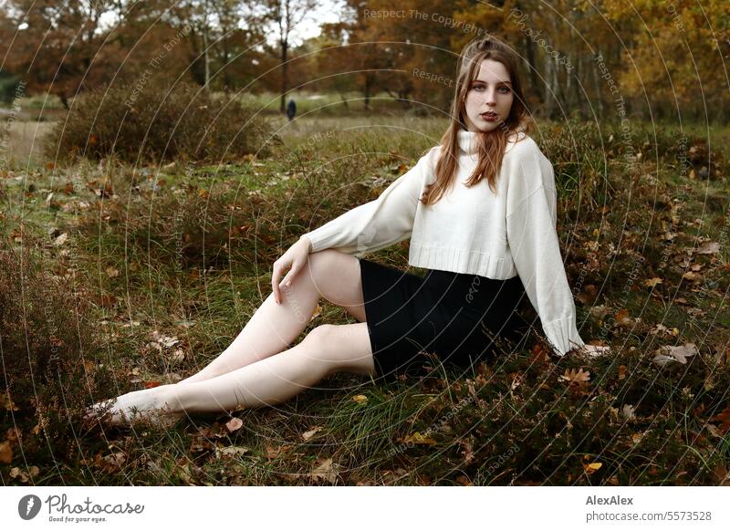 Young woman in a white wool sweater and black skirt sits barefoot in the grass among heather in a northern German heath landscape in the fall Woman Large