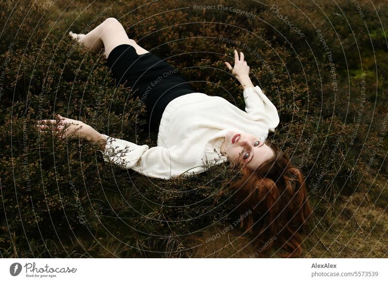 Young woman in white woolen sweater and black skirt lying upside down on a rampart in the heath between grass and heather Woman Large tall woman long legs Slim
