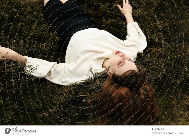 Young woman in white woolen sweater and black skirt lying upside down on a rampart in the heath between grass and heather Woman Large tall woman Slim Bright Joy