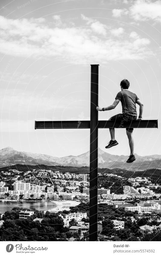 Young man sits on a cross and looks into the distance Belief Crucifix God religion Man Hope Spirituality willpower Symbols and metaphors Town mountain Rear view