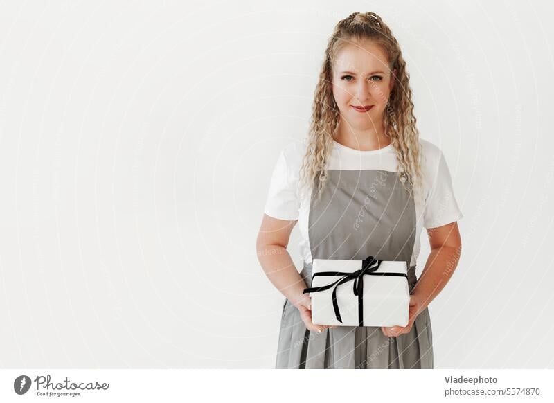 Young woman wearing apron hold in hand white present box with gift ribbon bow. Black white birthday surprise holiday holding event background celebration person