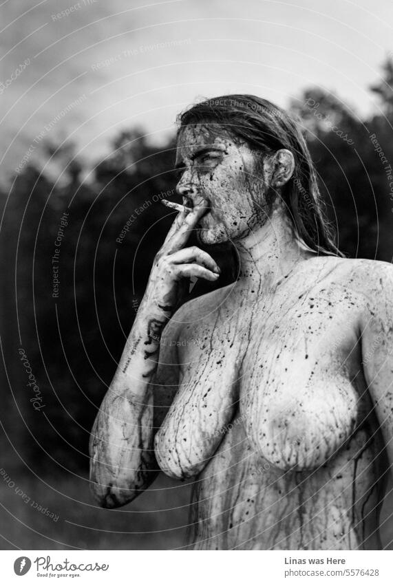 A black and white image with a naked and wild girl in it. She’s smoking a cigarette, showing her nude sexy curves, with some mud and body paint on her. A nude model is feeling wild and free in her own skin. And a bit devilish.