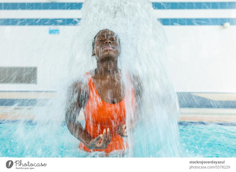 Black young woman massaging her back under a water jet at the swimming pool black person sports blue active female heated swimsuit orange healthy smiling