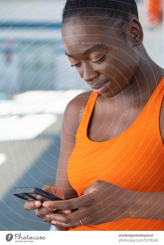 Black young woman using her smartphone, sitting in a bench at the swimming pool female women person lifestyle mobile black heated swimsuit orange healthy
