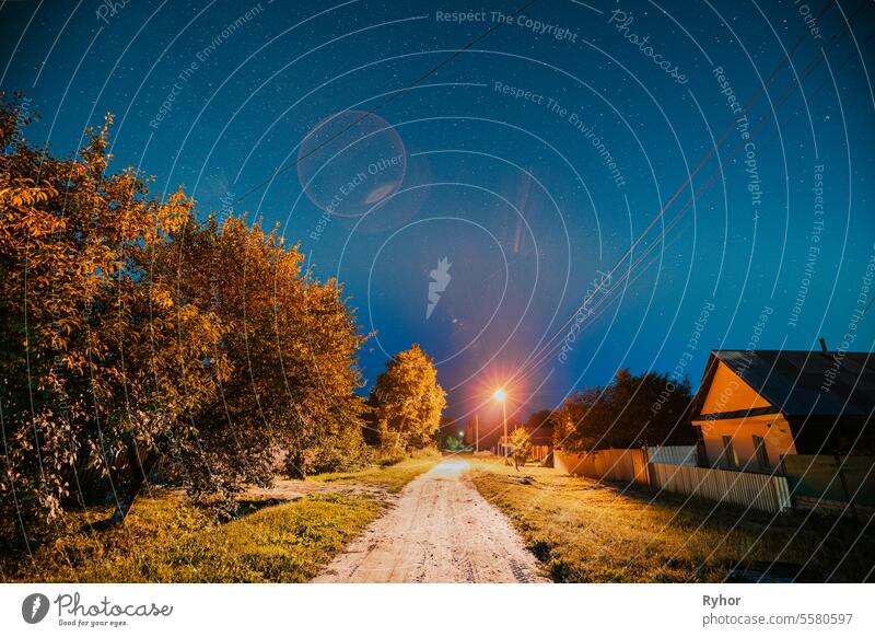 Night sky over street with houses in village. Night starry sky above village with bright stars and meteoric track trail. Glowing stars above summer nature