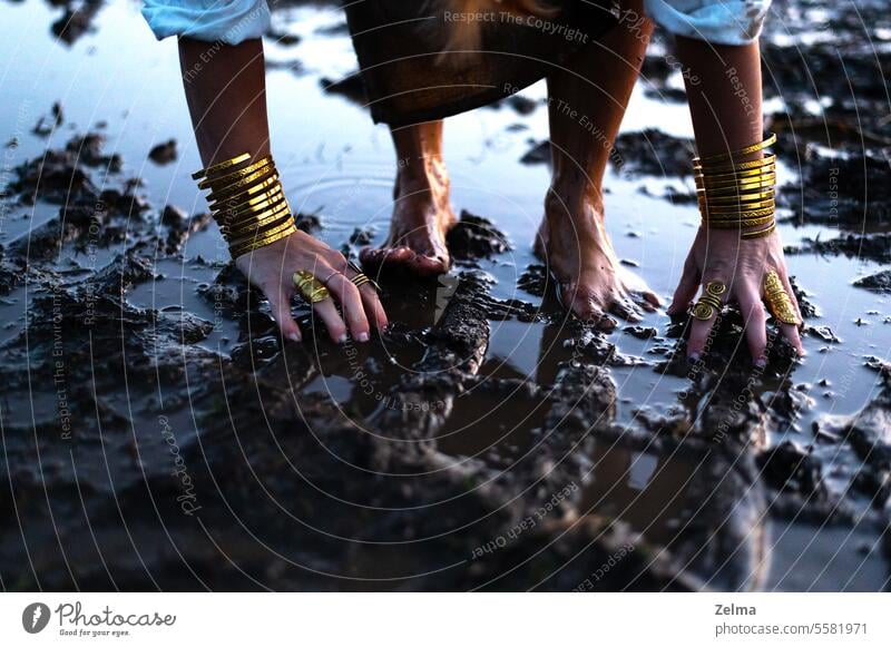 A woman's bare feet and hands with luxury bracelets and rings in a dirty puddle of black mud legs female jewelry golden muddy field escape barefeet family