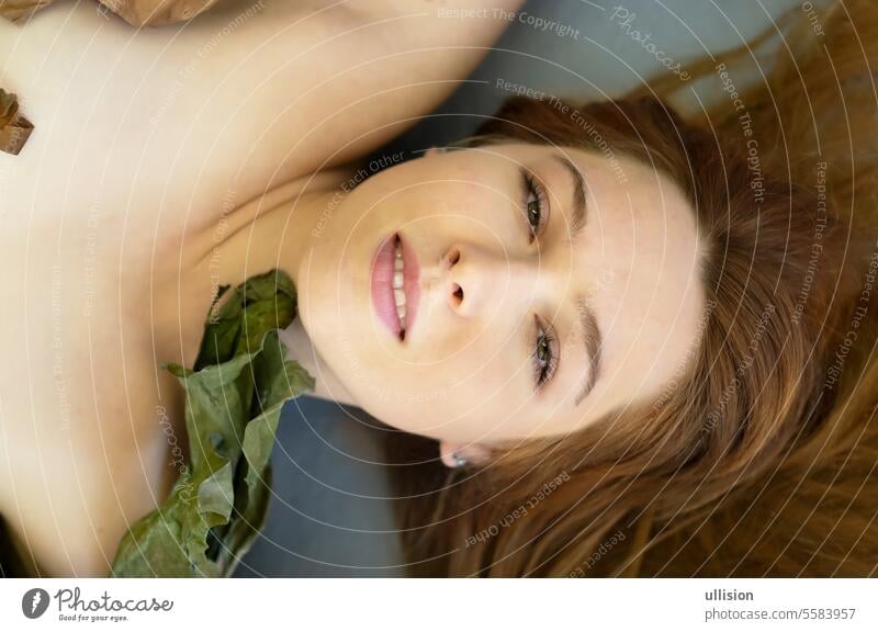sensual seductive portrait of a young redhead woman lying between green and dry plant leaves decorative copy space vogue hair banana tree gray studio floor