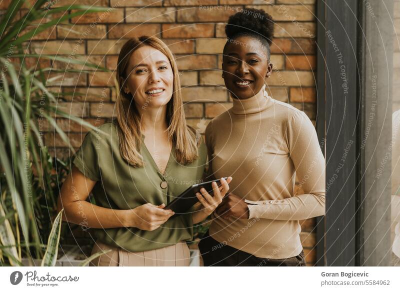 Two young business women with digital tablet standing by the brick wall in the industrial style office adult african american black businessman businesspeople