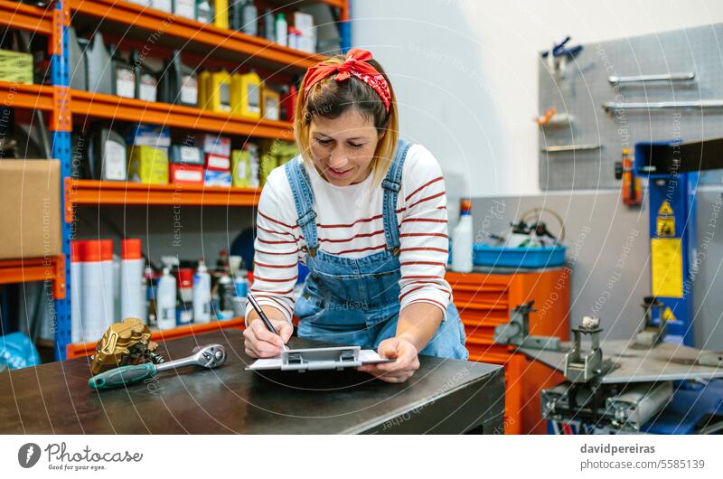 Woman checking stock in mechanic workshop woman female product merchandise garage store review information wear jeans overall bandana factory writing pen