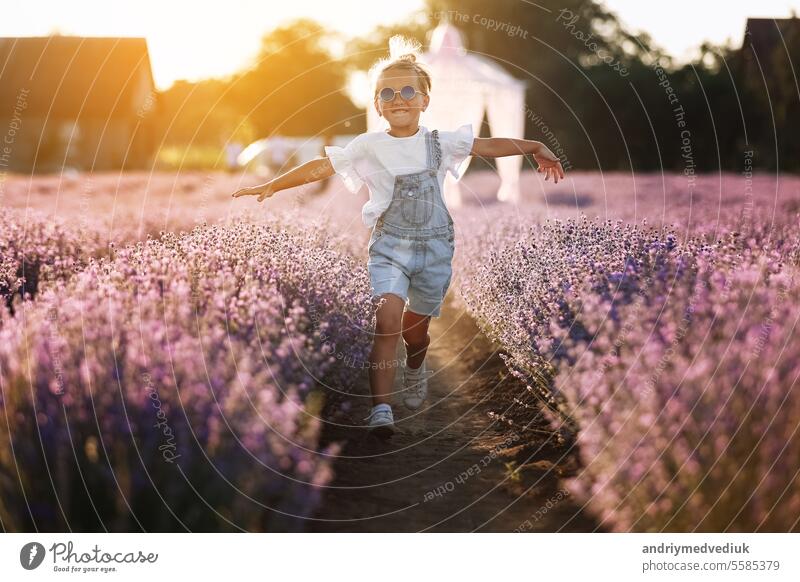 happy child girl is walking in lavender field on sunset. Smiling kid in suglasses , jeans jumpsuit is having fun in nature on summer day. Cheerful little girl. happy childhood concept