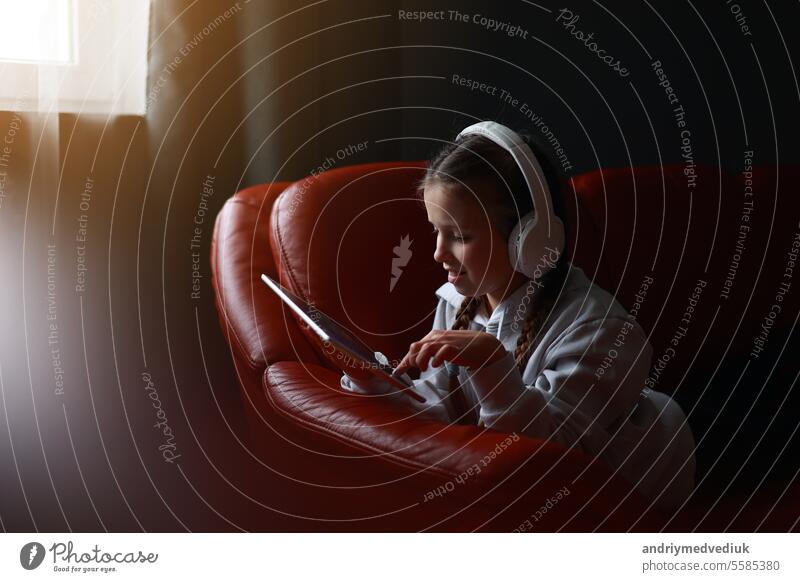 Concentrated teen girl in headphones plays game, communicate on internet social media, listens to music or online studies at home on digital tablet sitting on sofa. Children tech addiction concept