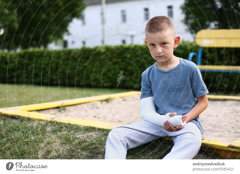 child caucasian with broken limb outdoors sits near the playground and looking on the plaster bandage on his arm. the worst summer vacation. concept of health accident, and medical