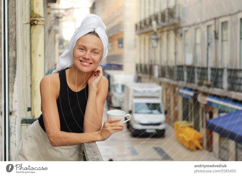 Young beautiful woman in a towel on head is drinking coffee or tea and smiling cute standing on balcony on ueropean street background. lifestyle morning. Concept of domestic or traveling lifestyle.
