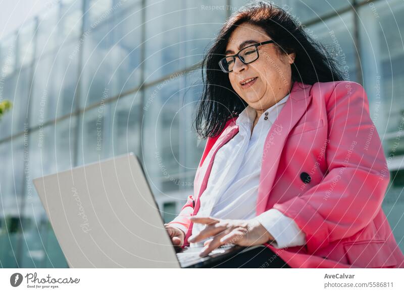 Smiling adult woman having an online meeting on laptop sitting on a bench in a place outside work. women female person computer technology business happy