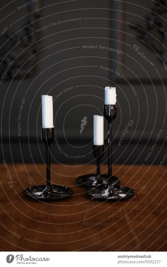 Modern three black glass candlesticks with white candles on wooden table in antique style on dark wall background. Minimalist elegant decor with candle holders with wax different size.