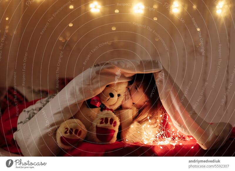 little cute smiling baby girl with white teddy bear peeking out from under a blanket on a bed at home with lighting garlands at dark. Child looking in camera. Fairytale childhood with miracle concept
