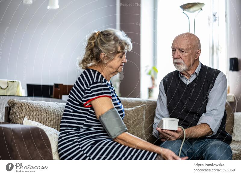 Senior couple measuring blood pressure while sitting on sofa in living room at home real people woman senior mature female together Caucasian elderly house old