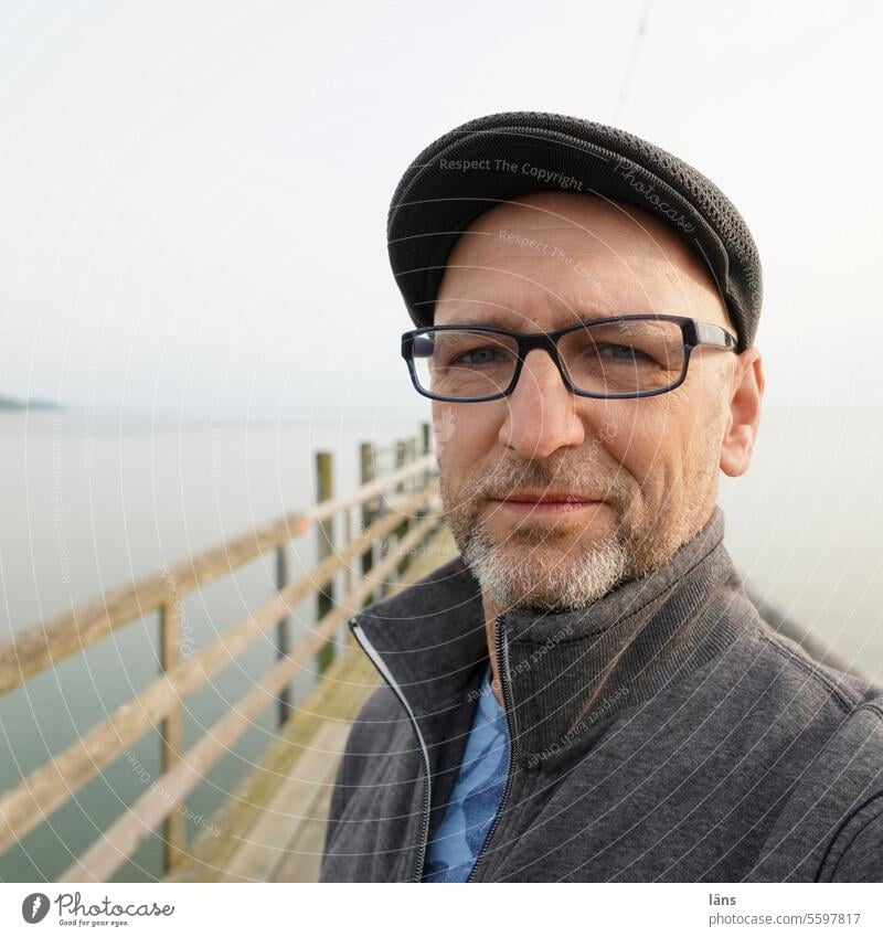 What comes what stays ll Man portrait Horizon standing Eyeglasses Cap Facial hair Face Looking into the camera Exterior shot on the lakeshore Contentment