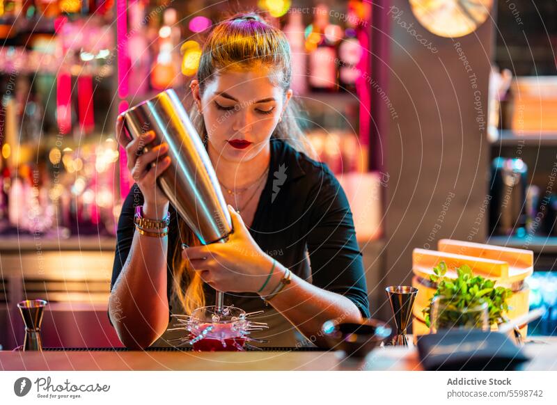 Focused young female barmaid with blonde hair standing at counter while making cocktail at illuminated bar woman pouring bottle glass alcohol prepare beverage