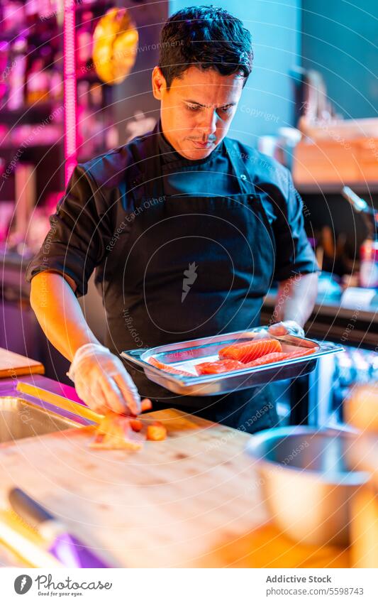 Focused Chef in black apron and gloves standing while preparing oriental dinner with salmon meat at counter at sushi bar man cook tray prepare meal illuminated