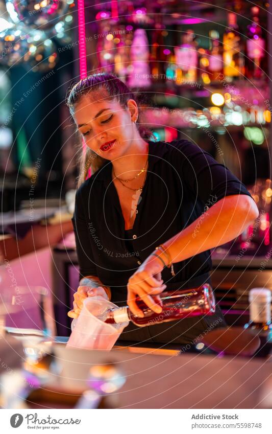 Smiling young Female barkeeper standing while pouring drink in mug from glass bottle at pub woman barmaid counter illuminated expertise skill focus positive