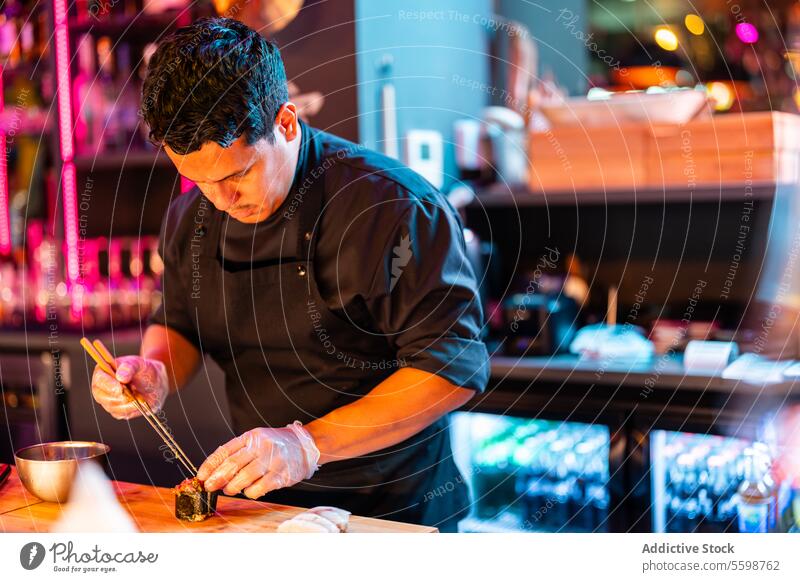 Focused Cook in black apron and gloves using chopsticks for making fresh sushi roll while working at sushi bar man chef prepare kitchen dinner oriental