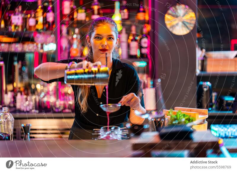 Beautiful barmaid with blonde hair looking at camera while preparing cocktail at pub woman barkeeper pouring shaker strainer confident young straining drink