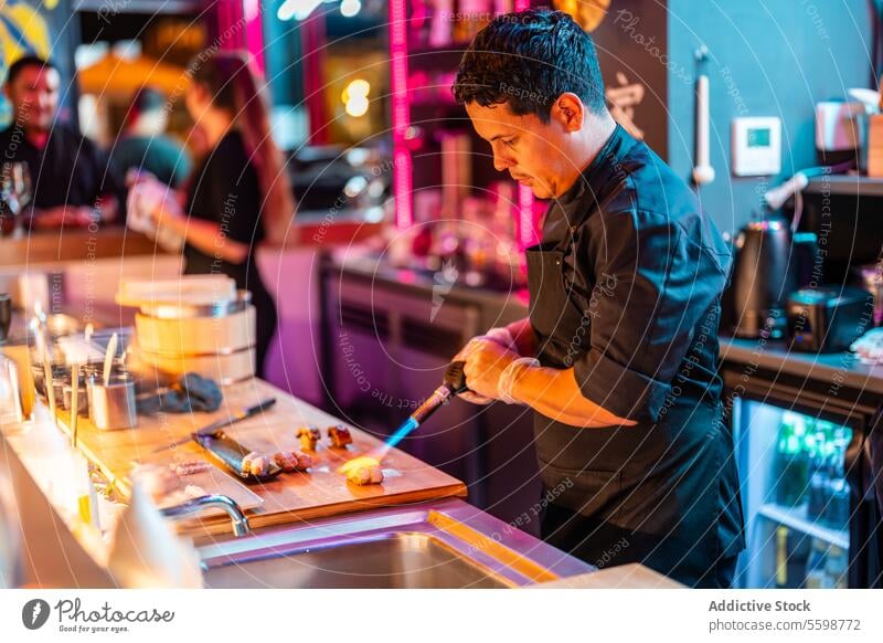 Focused male cook in black apron standing while using blow torch on fresh sushi at chopping board in modern kitchen man heat chef prepare oriental dinner