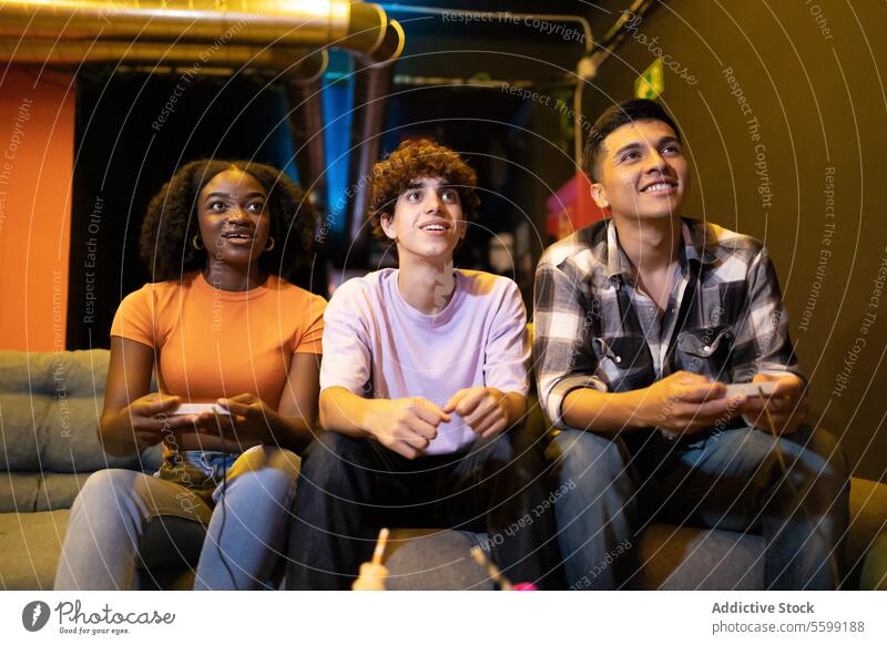 Three young multiracial friends are sitting on a couch engaged in a fun video game session men diverse entertainment multiethnic controller indoor living room