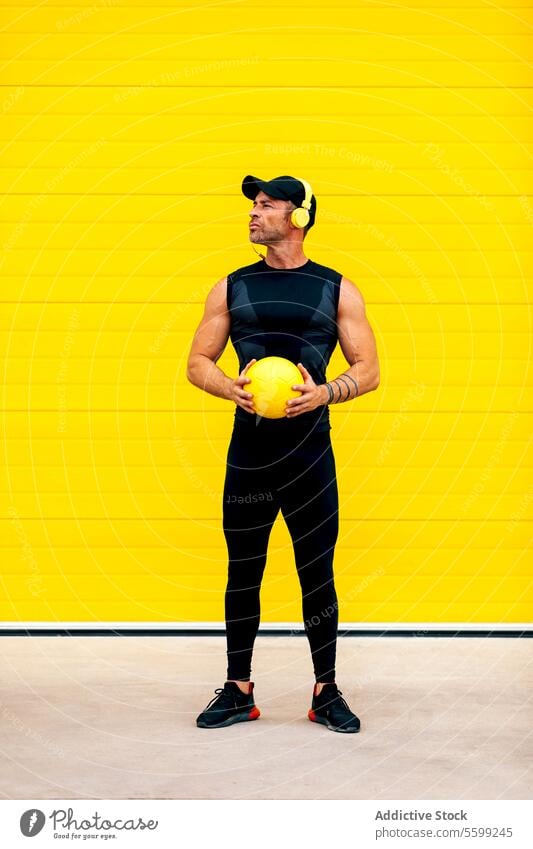 Confident sportsman with ball standing in city athlete headphones looking away confident muscular holding street wall serious play game half face cap activewear