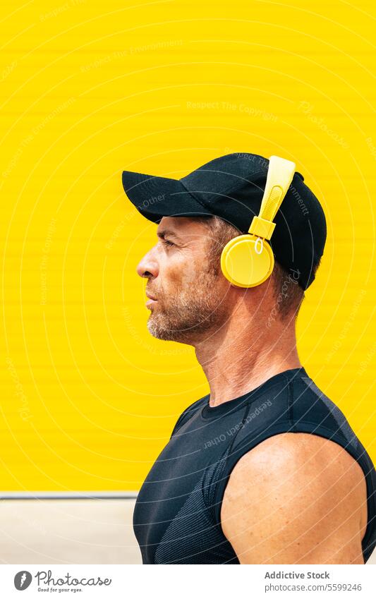 Closeup side view of serene mid adult male athlete in black cap and sportswear looking away while listening music through headphones against yellow background