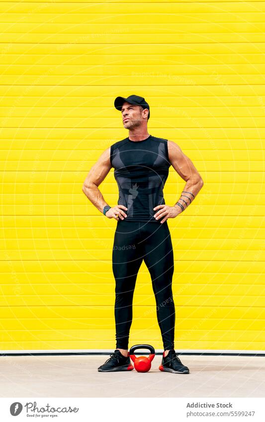 Full body of sportsman standing in city during break athlete kettlebell arms akimbo rest muscular street yellow wall training cap activewear weightlifting