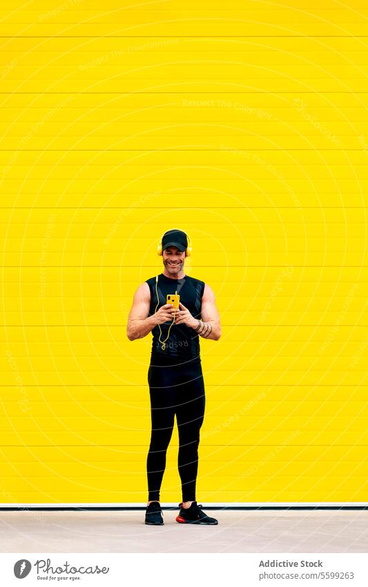Confident male athlete using phone during workout smartphone smile listen music headphones street yellow wall full body cap confident happy standing city
