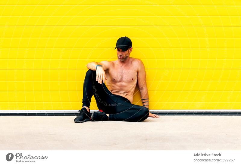 Tired shirtless athlete sitting on street sportsman naked torso exhausted muscular think workout active tired full body relax yellow wall cap rest contemporary