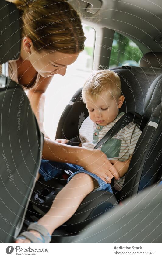Happy mother fastening seat belts of cute kid son sitting in car seat journey countryside summer female young boy child mom parent vehicle tree transport nature