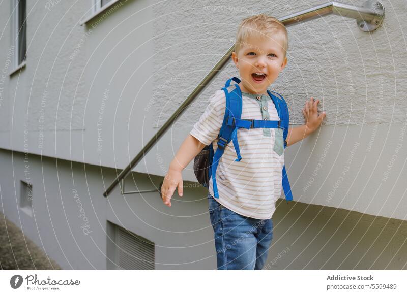 Happy cute kid boy with backpack standing near gray wall building in daylight child smile railing cheerful childhood playful path happy summer adorable