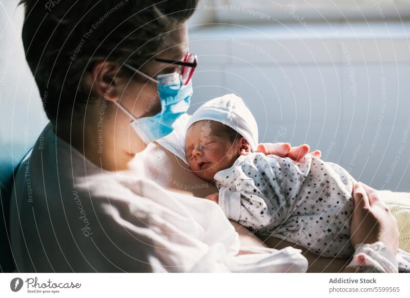 Mother wearing a face mask while holding her newborn baby in the hospital room. women mother beginnings parent bonding affectionate connection joy enjoyment