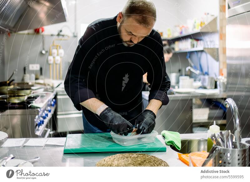 Bearded male professional chef in black gloves arraigning food into transparent container while working in professional kitchen man restaurant prepare colleague